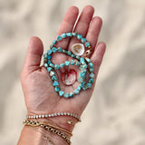 A hand holding an Endless Summer Necklace - Sea + Sky with two shells, and wearing several bracelets, including a Love Set Match Bracelet, a Diamond Cut Bracelet, a Rolo Bracelet and an Element Short Link Bracelet with a blurred sand background. 