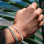 Close-up of a handing wearing two Endless Summer Bracelets (Turqs + Caicos and Blue Lagoon) with palm leaves in the background. 