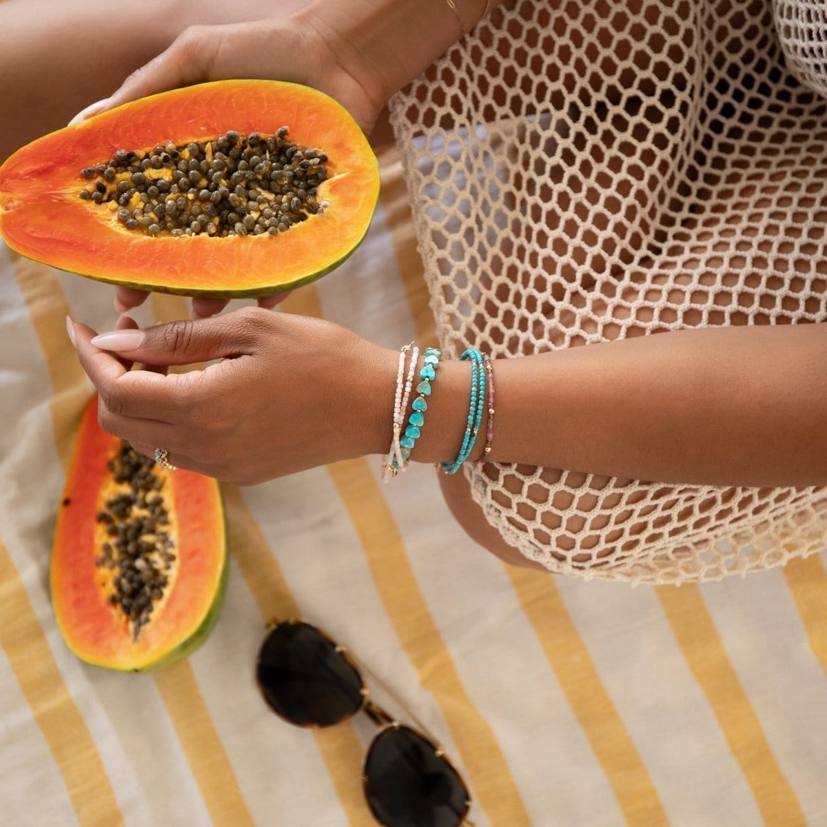 A hand wearing Endless Summer Bracelets in Coral Reef, Sea + Sky, Pink Sands and Turqs + Caicos, holding a papaya in a cream mesh dress, with another papaya and sunglasses placed on a yellow and white striped background.