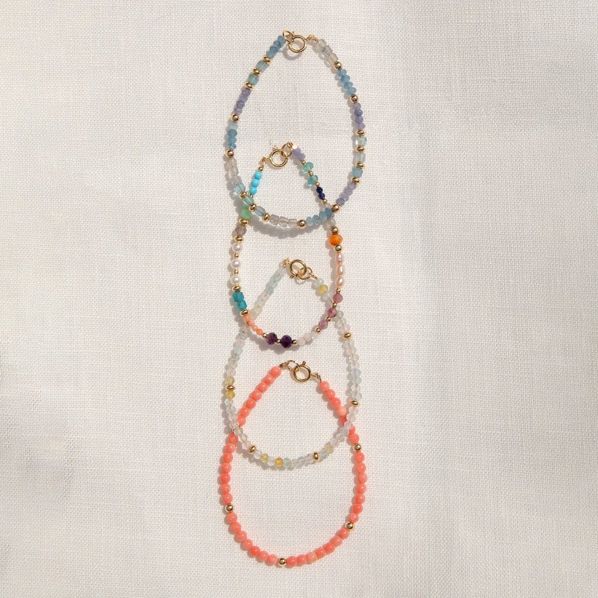 Four Endless Summer Bracelets (Blue Lagoon, Capri, Mykonos and Saint-Tropez) laid vertically on an off-white fabric background. 