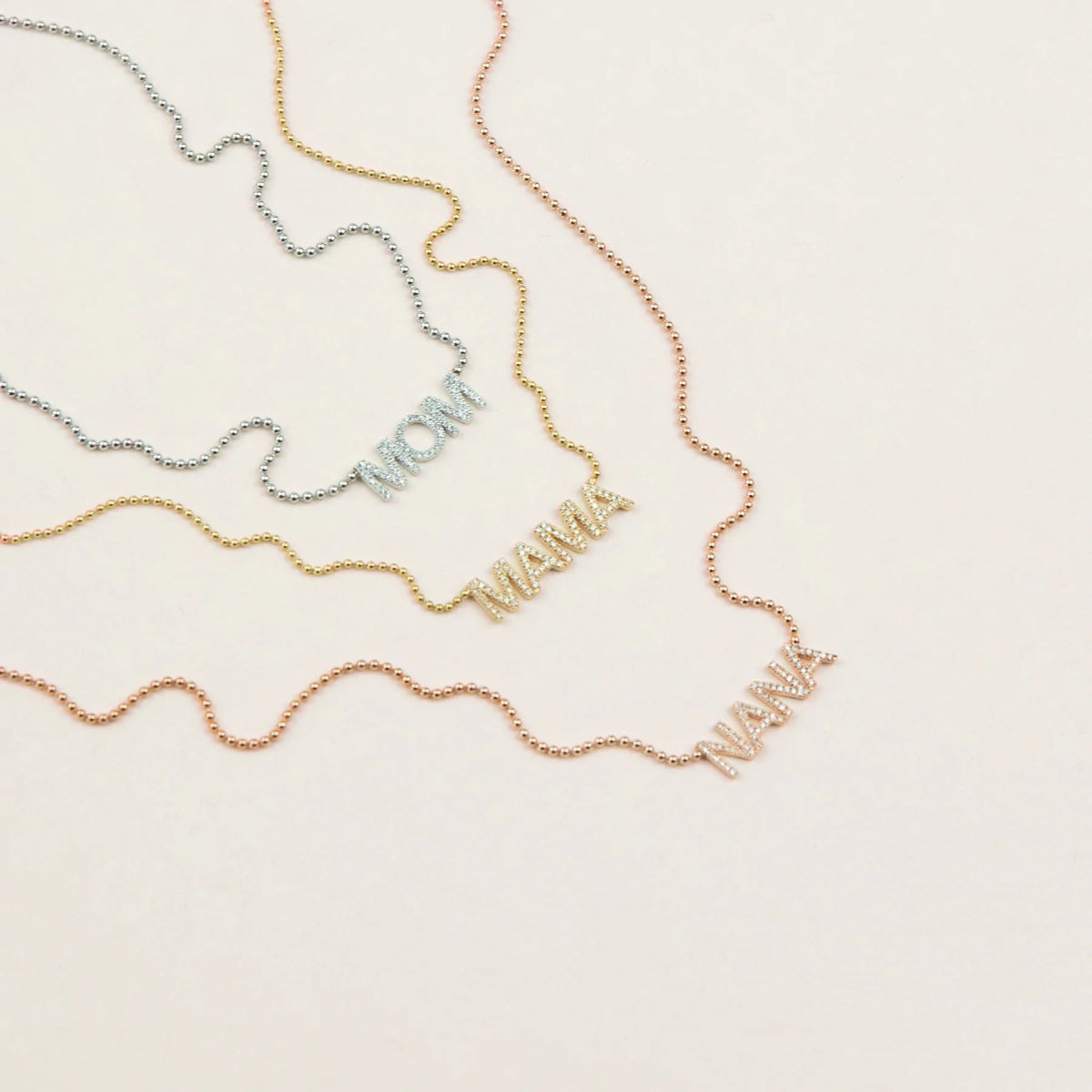 A Pavé MOM Necklace in white gold, a Pavé MAMA Necklace in yellow gold and a Pavé NANA Necklace in rose gold, in a cream background. 