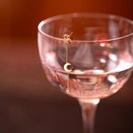 A Luna Spritz Earring on top of a cocktail glass against a blurred red background. 