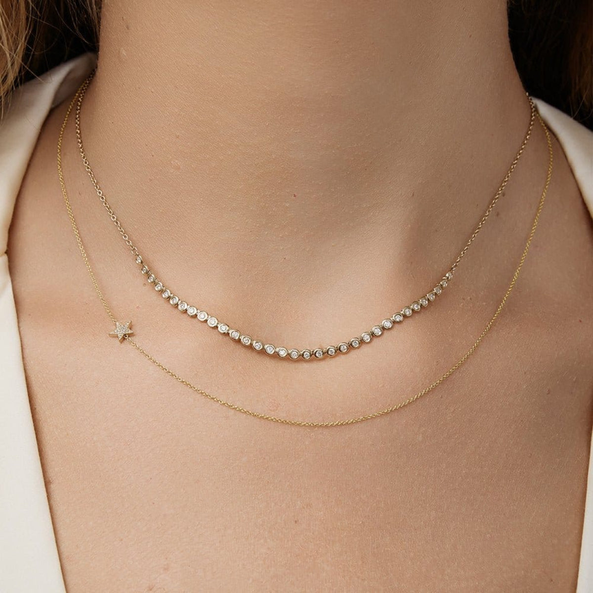 Close-up of a neck wearing The Everyday Diamond Necklace layered with a Custom Gold Necklace - 1 Letter with Pave Star, both in yellow gold, with a white top.