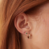 Close-up of a 14K Gold Pavé Letter Stud in J, a Birthstone Stud - Emerald (May), and a Gold Huggie worn in a woman’s ear, with orange hair visible.