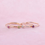 Three delicate rose gold rings with small colorful gemstones, displayed on a soft pink background.