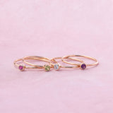 Three delicate rose gold rings with small colorful gemstones, displayed on a soft pink background.