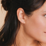 Close-up of a yellow gold Birthstone Hoop and Birthstone Stud in a brunette woman's ear against a neutral background. 