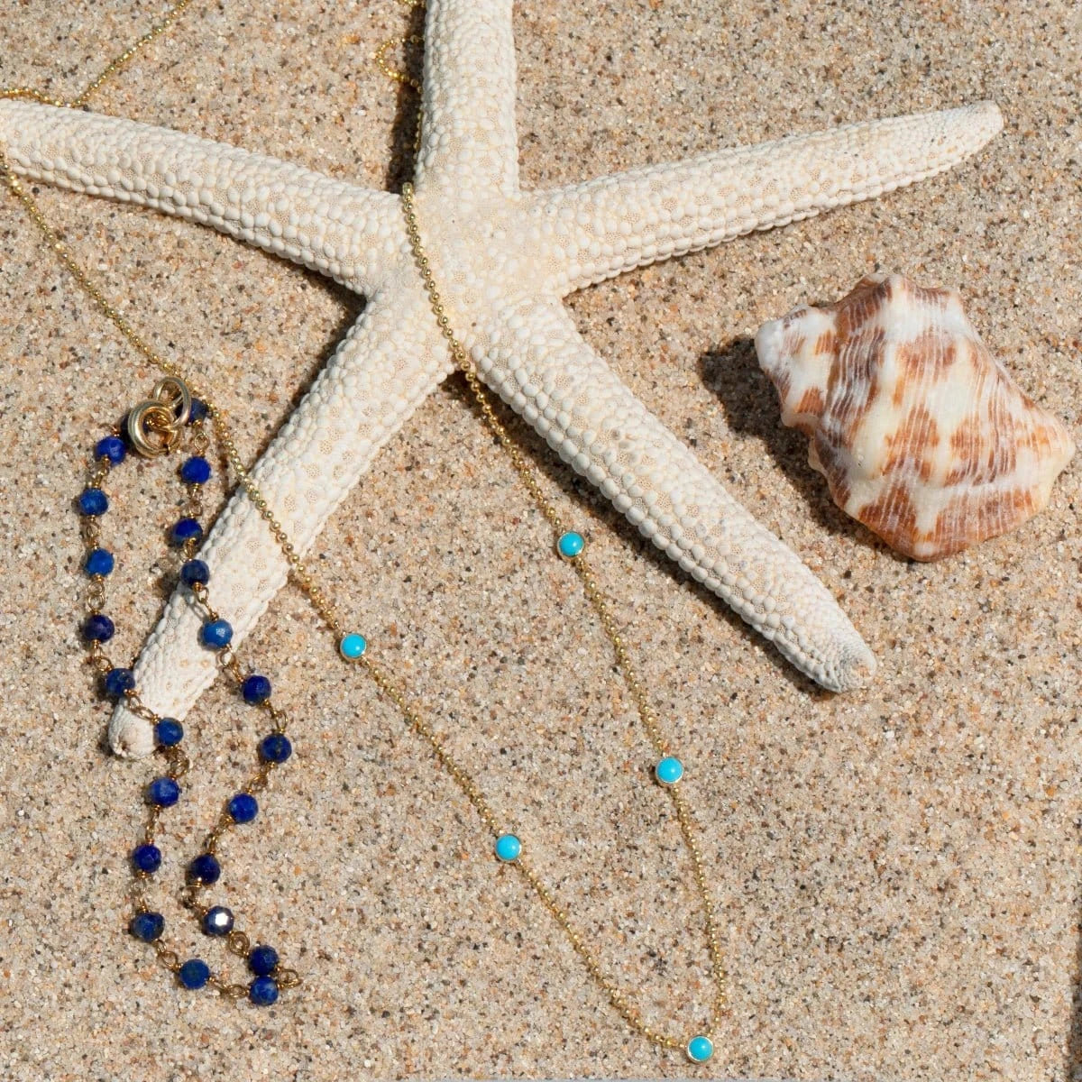 The Thistle Bouquet Bracelet and Quintet Birthstone Layering Necklace in yellow gold with turquoise stones, both laid in front of a starfish next to a shell against a sandy background.