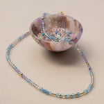 An Endless Summer Necklace - Blue Lagoon laid on top of a small coloured bowl with different coloured beads in it, against a light background.