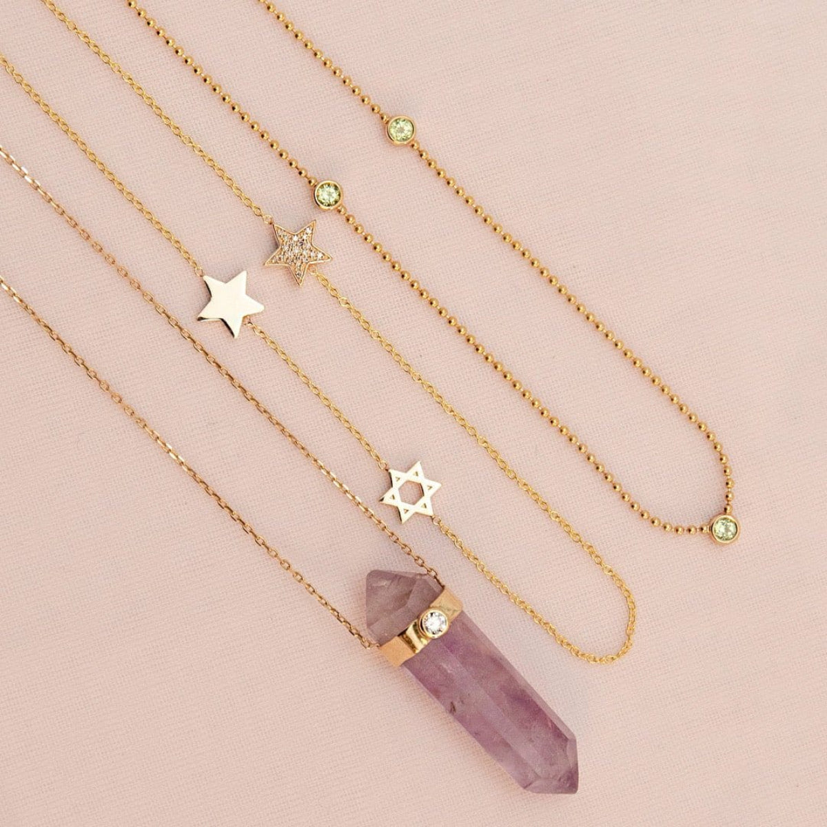 The Healing Retreat Necklace, a Custom Gold Necklace - 3 Letters with star charms and a Triple Birthstone Layering Necklace - Yellow Gold (Peridot) laid on a light pink background. 