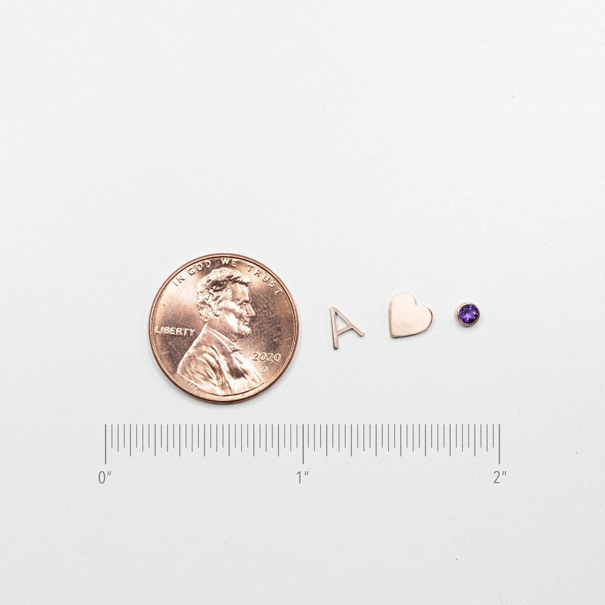 A coin next to an A, heart and birthstone charm with a ruler underneath showing the size of the charms.
