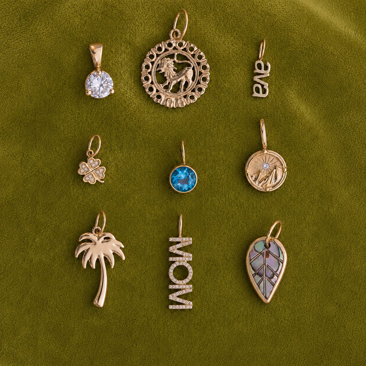 Nine gold-toned charms and pendants arranged in a 3x3 grid on an olive green textured fabric background. The charms include a round clear stone, a circular animal motif, the letters 'ANA', a clover, a blue gemstone, a circular bird motif, a palm tree, the word 'MOM', and a leaf with inlay details.