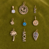 Nine gold-toned charms and pendants arranged in a 3x3 grid on an olive green textured fabric background. The charms include a round clear stone, a circular animal motif, the letters 'ANA', a clover, a blue gemstone, a circular bird motif, a palm tree, the word 'MOM', and a leaf with inlay details.