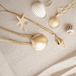 A Chic Cuban Curb Chain with The Golden Venus Shell, Element Long Link Chain with a Golden Scallop Shell and a Golden Spiral Shell on The Slide Chain against a sandy background. 