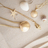 A Chic Cuban Curb Chain with The Golden Venus Shell, Element Long Link Chain with a Golden Scallop Shell and a Golden Spiral Shell on The Slide Chain against a sandy background. 