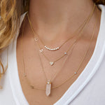 A close-up of a woman’s neck wearing four yellow gold necklaces: two Custom Gold Necklaces (one with three charms and the other with four charms), the Clarity Retreat Necklace and the Diamond Arc Layering Necklace, with a cream top.
