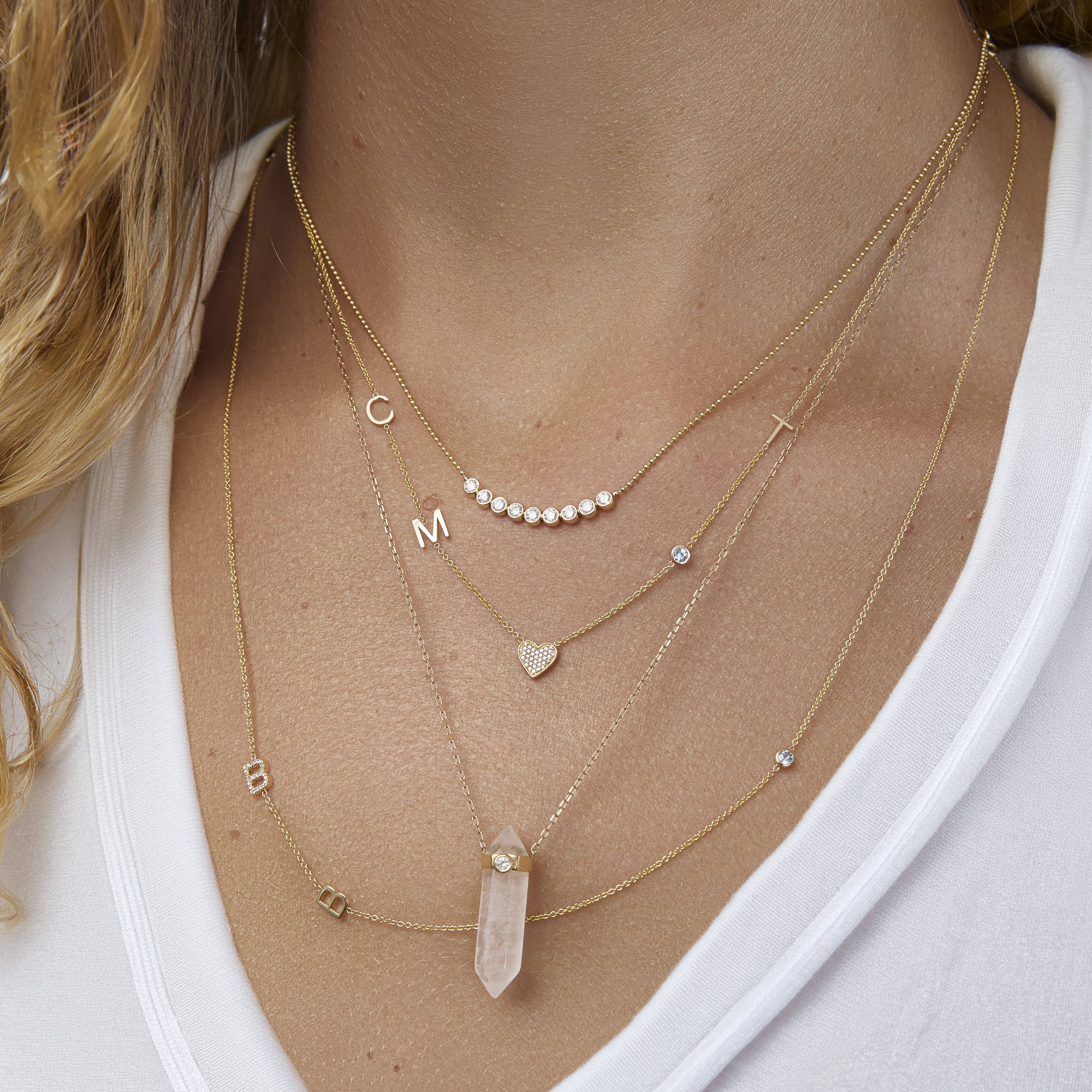 A close-up of a woman’s neck wearing four yellow gold necklaces: two Custom Gold Necklaces (one with three charms and the other with four charms), the Clarity Retreat Necklace and the Diamond Arc Layering Necklace, with a cream top.