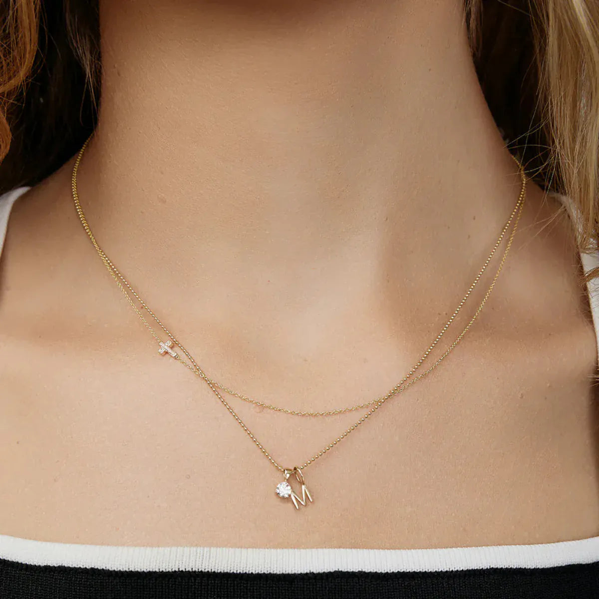 A woman's neck wearing a yellow gold Classic Bead Chain with a Gold Letter Jumble - M, and a 14k Gold Asymmetrical Charm Necklace - Pave Cross. 