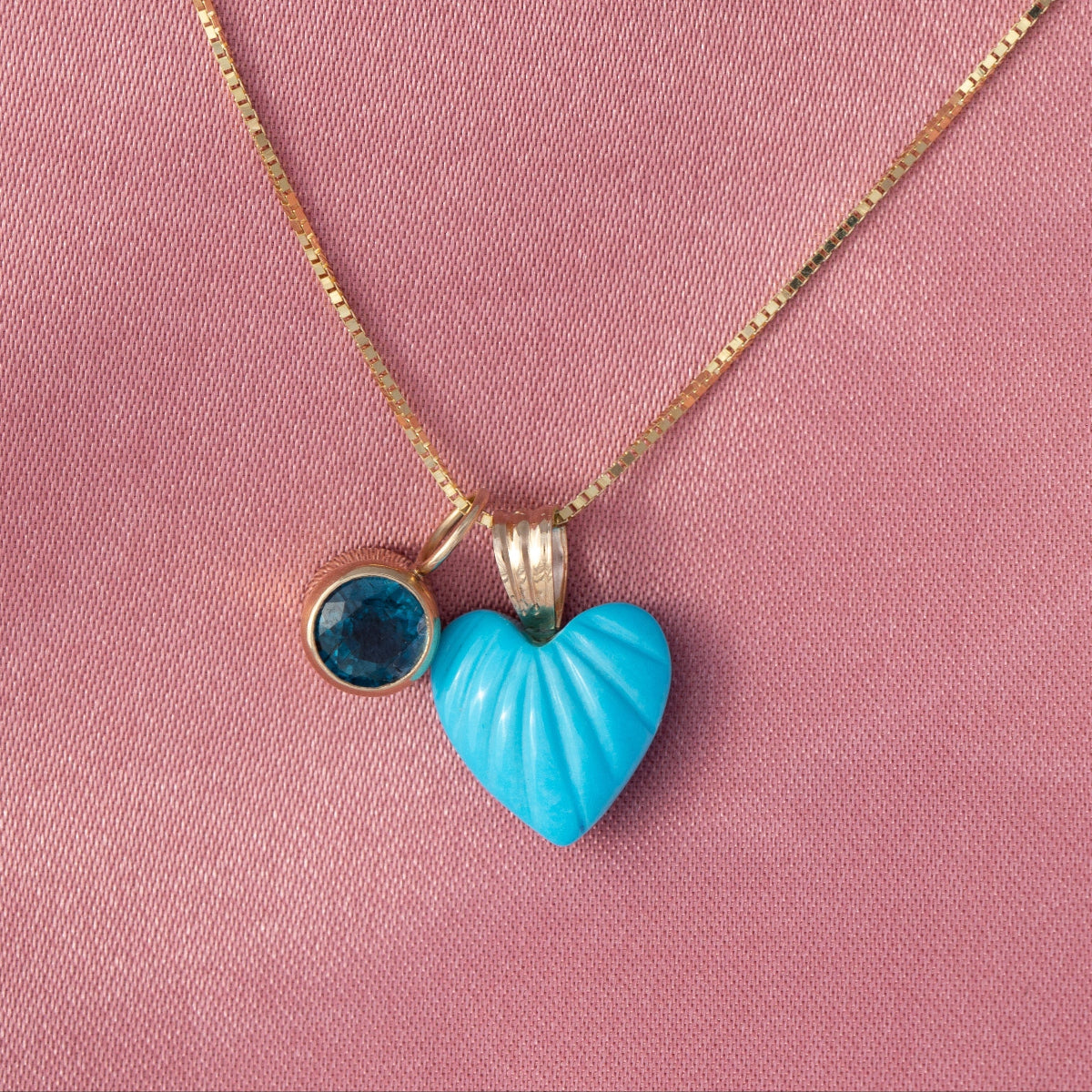 An Etched Turquoise Heart Charm and a Color Pop Pendant in Blue (London Topaz) on a Box Chain against a pink background. 