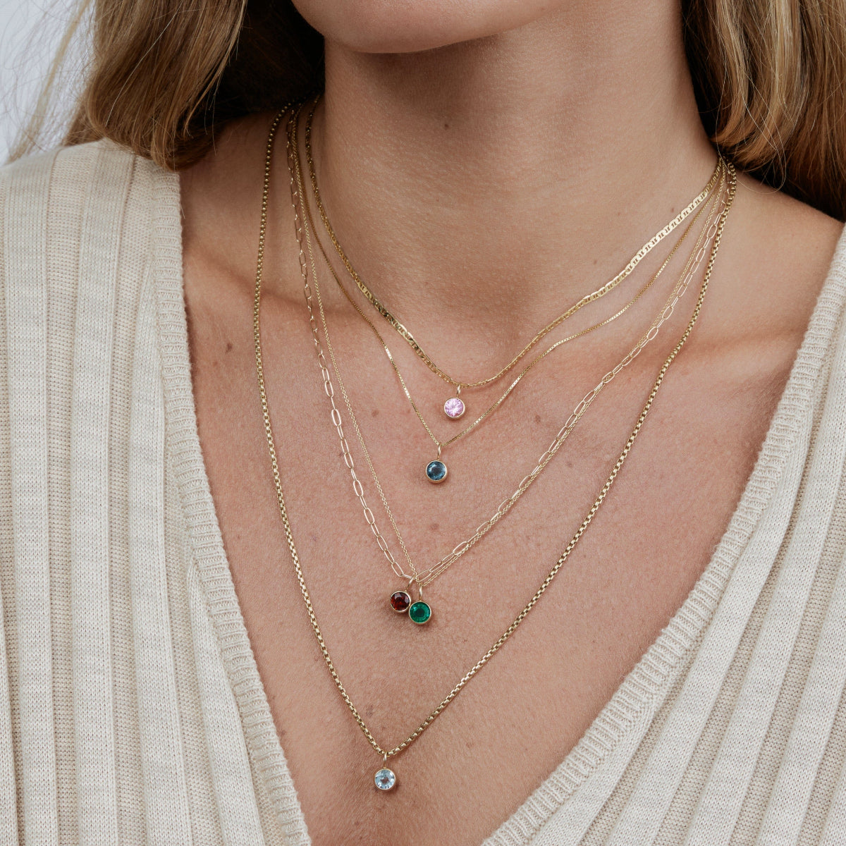 Close-up of a woman wearing an Overboard Layering Chain with a Pink (Pink Tourmaline) Color Pop Pendant, The Slide Chain with a Blue (London Topaz) Color Pop Pendant, an Element Short Link Chain with a Red (Garnet)Color Pop Pendant, Signature Cable Chain with a Green (Lab Grown Emerald) Color Pop Pendant and a Box Chain with a Light Blue (Aquamarine), styled in a cream top. 