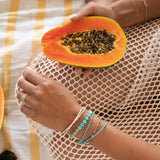 A hand wearing several Endless Summer Bracelets (Pink Sands, Turqs + Caicos, Coral Reef and Sea + Sky) and two Bouquet Rings, holding a papaya in the other hand with an Element Short Link Bracelet, styled in a cream mesh dress with a white and yellow striped background.