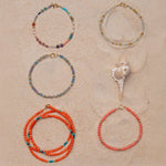 Four Endless Summer Bracelets (Capri, Saint-Tropez, Blue Lagoon and Mykonos) and an Endless Summer Necklace (Calypso) on a sandy background with a shell. 