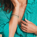 A close-up of a woman wearing multiple Bouquet Bracelets in different colours, a Classic Bead Chain, a Diamond Arc Layering Necklace, a 14k Gold Asymmetrical Moon Charm Necklace, a Heartfelt Ring, a Delicate Band and several Birthstone Rings, styled in a turquoise blue top.