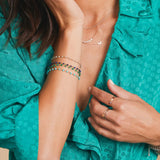 A close-up of a woman wearing multiple Bouquet Bracelets in different colours, a Classic Bead Chain, a Diamond Arc Layering Necklace, a 14k Gold Asymmetrical Moon Charm Necklace, a Heartfelt Ring, a Delicate Band and several Birthstone Rings, styled in a turquoise blue top.