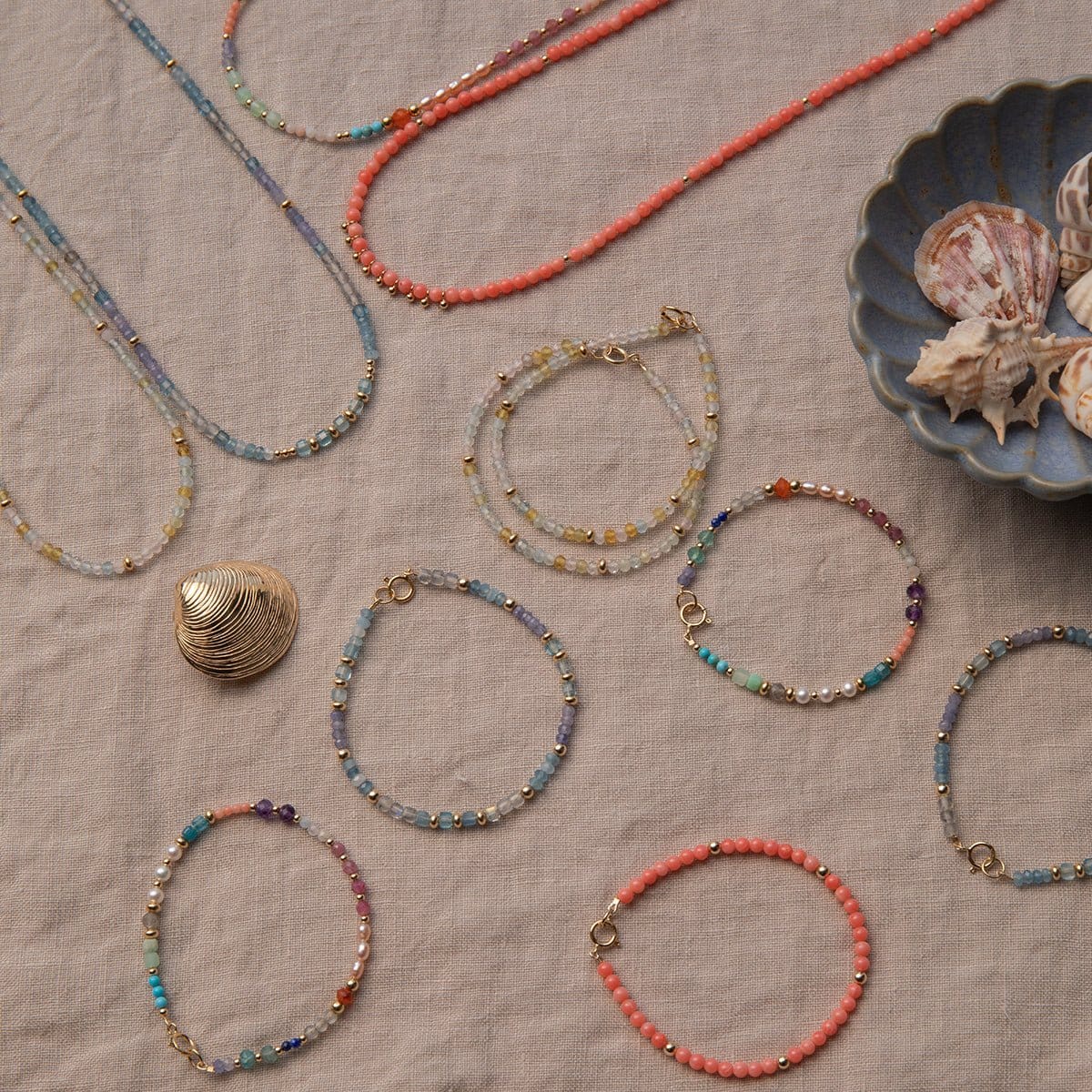 A range of Endless Summer Necklaces and Bracelets (Blue Lagoon, Mykonos, Saint-Tropez and Capri) laid on a beige fabric background with shells in a blue shell bowl. 