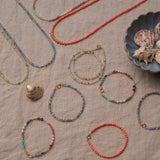A range of Endless Summer Necklaces and Bracelets (Blue Lagoon, Mykonos, Saint-Tropez and Capri) laid on a beige fabric background with shells in a blue shell bowl. 