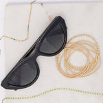 Two Confetti Necklaces in blue and green, bubble bracelets and black sunglasses on a light marble surface. 