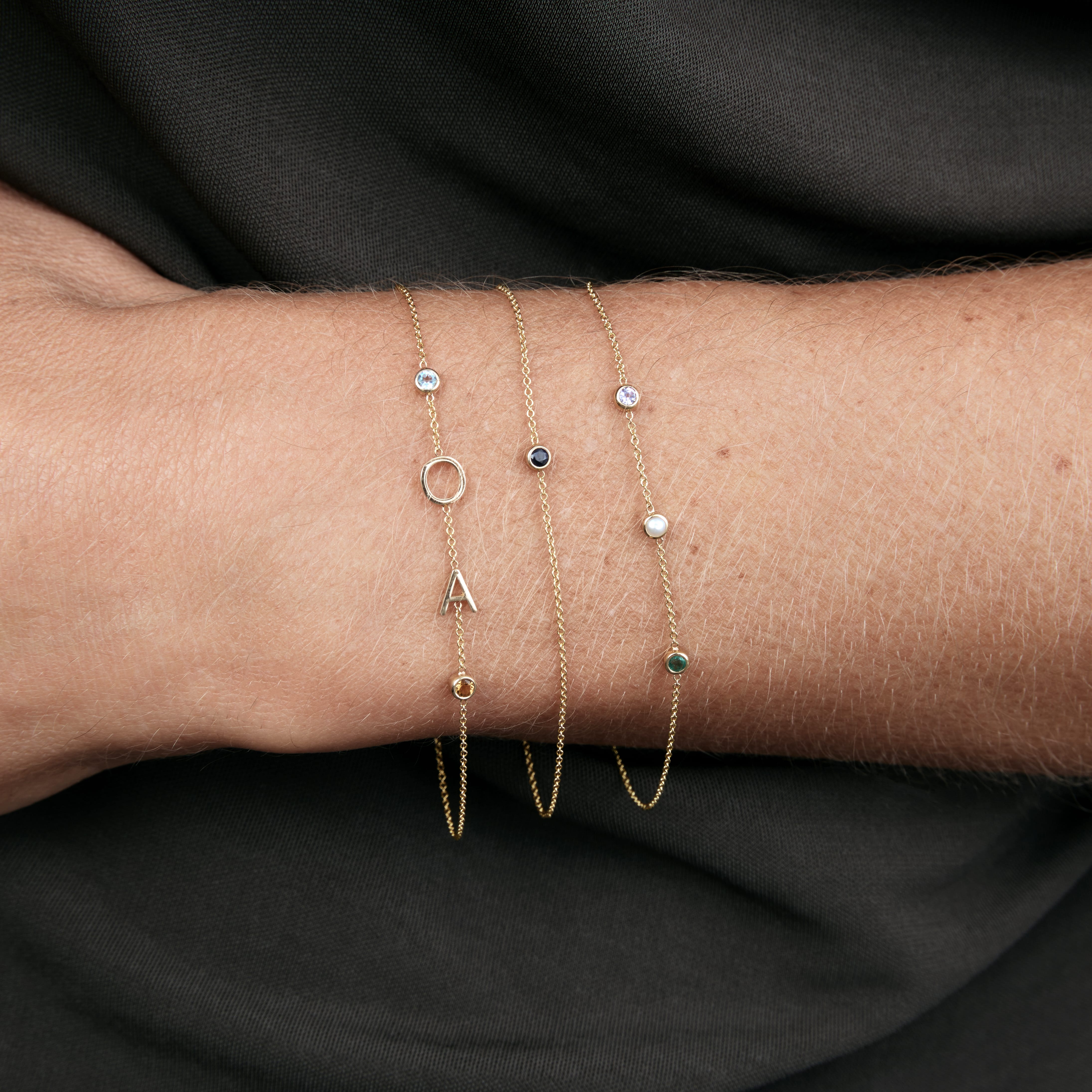 Three delicate gold chain bracelets with small gemstone and letter charms are shown worn on a person's wrist against a dark fabric background.