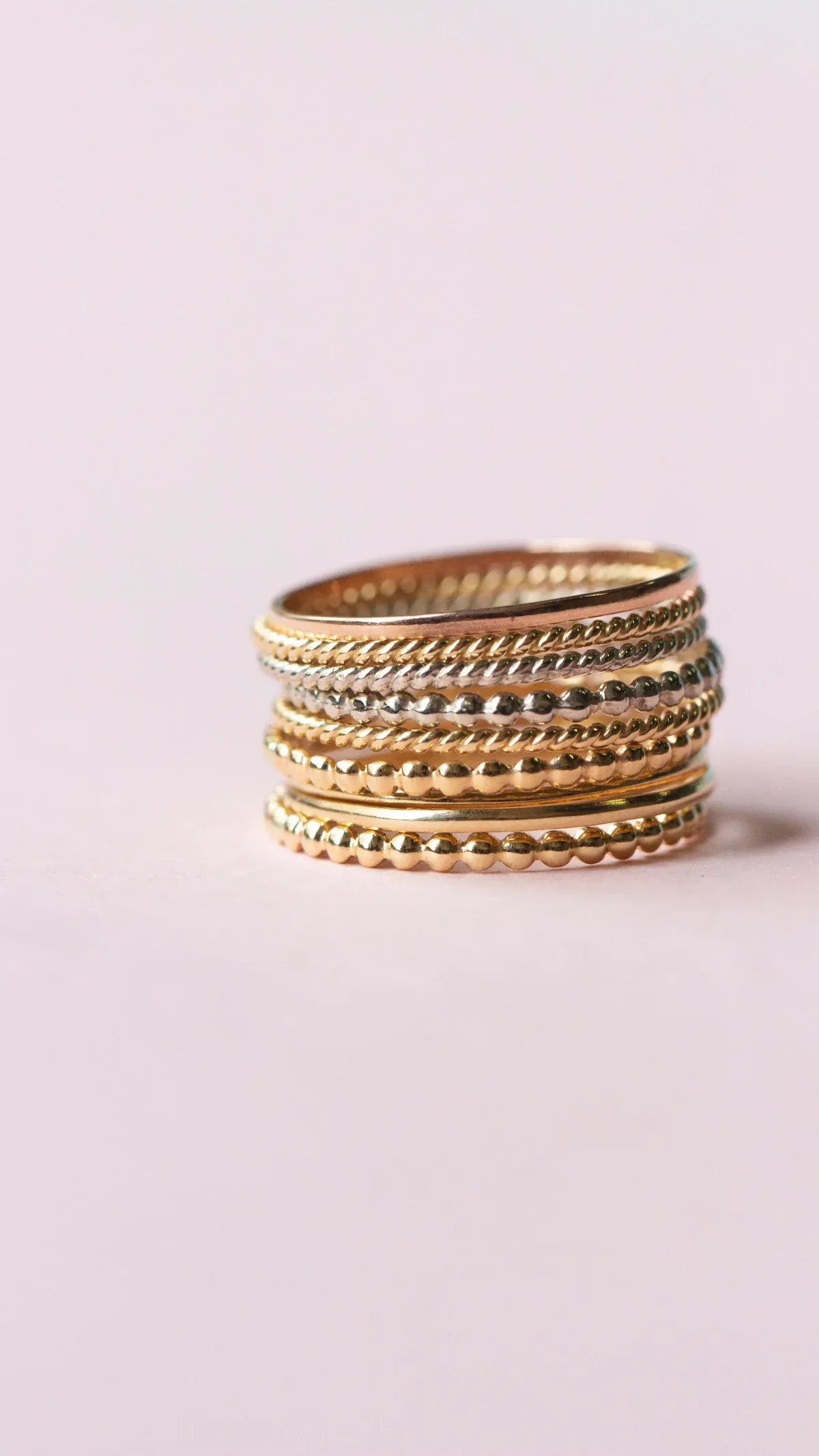 Several Delicate Trio Stacking Rings in yellow gold and white gold on a light pink background. 