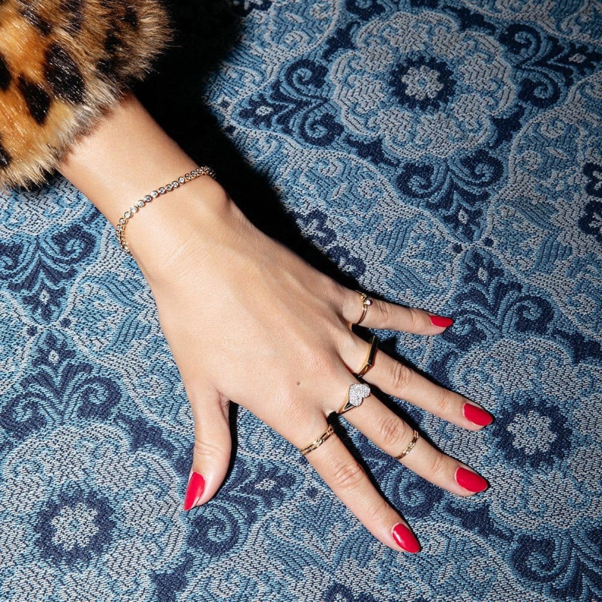 A hand wearing a Love Set Match Bracelet, a Link Ring, a Love on the Rocks Ring, a Geo Stacking Ring Square and two Birthstone Rings with red nails on top of a blue patterned fabric surface. 