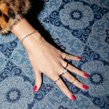 A hand wearing a Love Set Match Bracelet, a Link Ring, a Love on the Rocks Ring, a Geo Stacking Ring Square and two Birthstone Rings with red nails on top of a blue patterned fabric surface. 