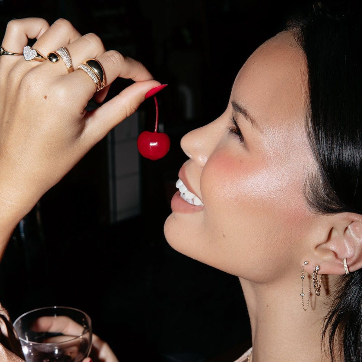 Close-up of a woman wearing a Diamond Fizz Earring, a Highball Diamond Chain Earring, two Pave Ear Cuffs, a Pinky Promise Ring, Love on the Rocks Ring, Make it a Double Ring, Cosmo Ring and Martini Ring, holding a cherry and a cocktail, against a dark background. 