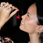 Close-up of a woman wearing a Diamond Fizz Earring, a Highball Diamond Chain Earring, two Pave Ear Cuffs, a Pinky Promise Ring, Love on the Rocks Ring, Make it a Double Ring, Cosmo Ring and Martini Ring, holding a cherry and a cocktail, against a dark background. 