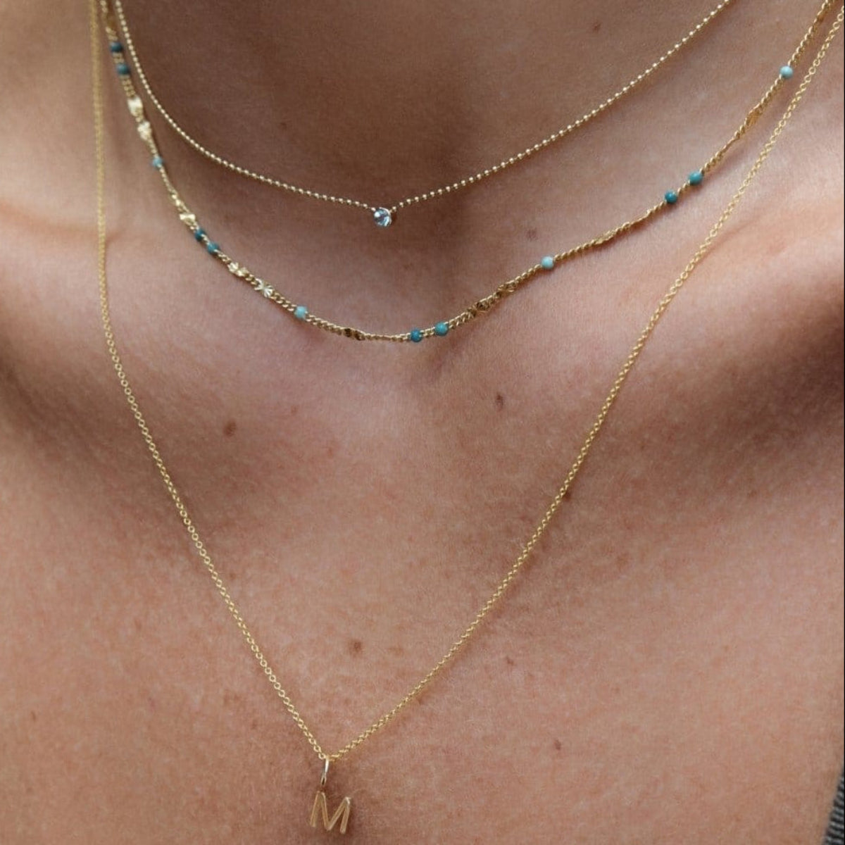 Close-up of a woman's collarbone area wearing a yellow gold Signature Cable Chain with a Gold Letter Charm in M, Confetti Necklace - Blue and a Single Diamond Layering Necklace. 