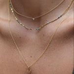 Close-up of a woman's collarbone area wearing a yellow gold Signature Cable Chain with a Gold Letter Charm in M, Confetti Necklace - Blue and a Single Diamond Layering Necklace. 