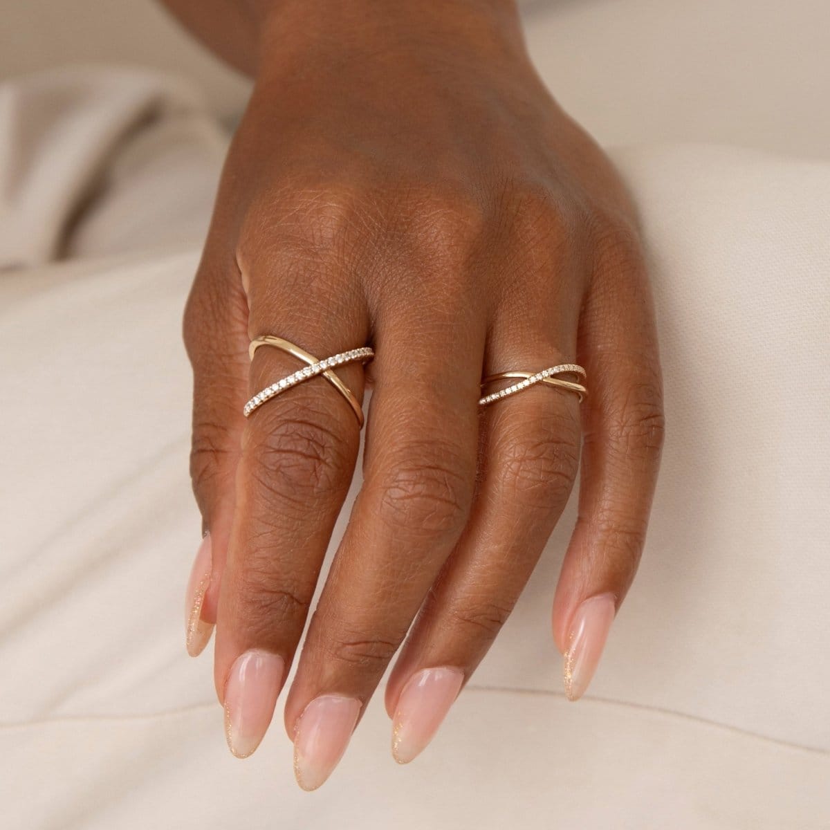 A hand wearing a yellow gold Criss-Cross Diamond Duo Ring and Criss-Cross Diamond Daze Ring with light pink nails against an off-white background. 