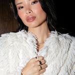 A woman wearing a Diamond Cascade Earring, The Ultimate Diamond Necklace, a Make it a Double Ring, Martini Ring, Cosmo Ring and Pinky Promise Ring, with cream fluffy jacket against a dark background. 