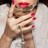 A hand wearing a C’est Moi Link Ring, a Make It a Double Ring and two Bouquet Rings, with red nails, holding a martini cocktail, styled with red lipstick and a cream fur jacket.
