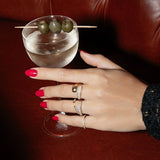 A hand wearing a Make it a Double Ring, Cosmo Ring, Open Diamond Ring and two Pave Diamond Rings with red nails holding a martini cocktail, against a brown leather background. 