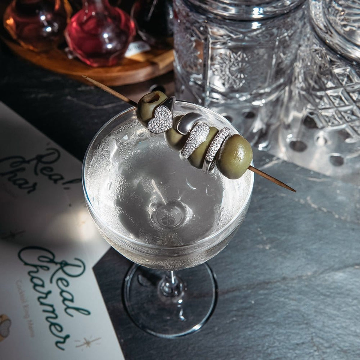 A white gold Love on the Rocks Ring, Make it a Double Ring and Cosmo Ring on a toothpick with green olives, on top of a cocktail glass, against a dark blue surface with other glasses. 