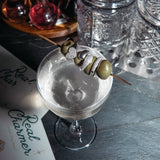 A white gold Love on the Rocks Ring, Make it a Double Ring and Cosmo Ring on a toothpick with green olives, on top of a cocktail glass, against a dark blue surface with other glasses. 