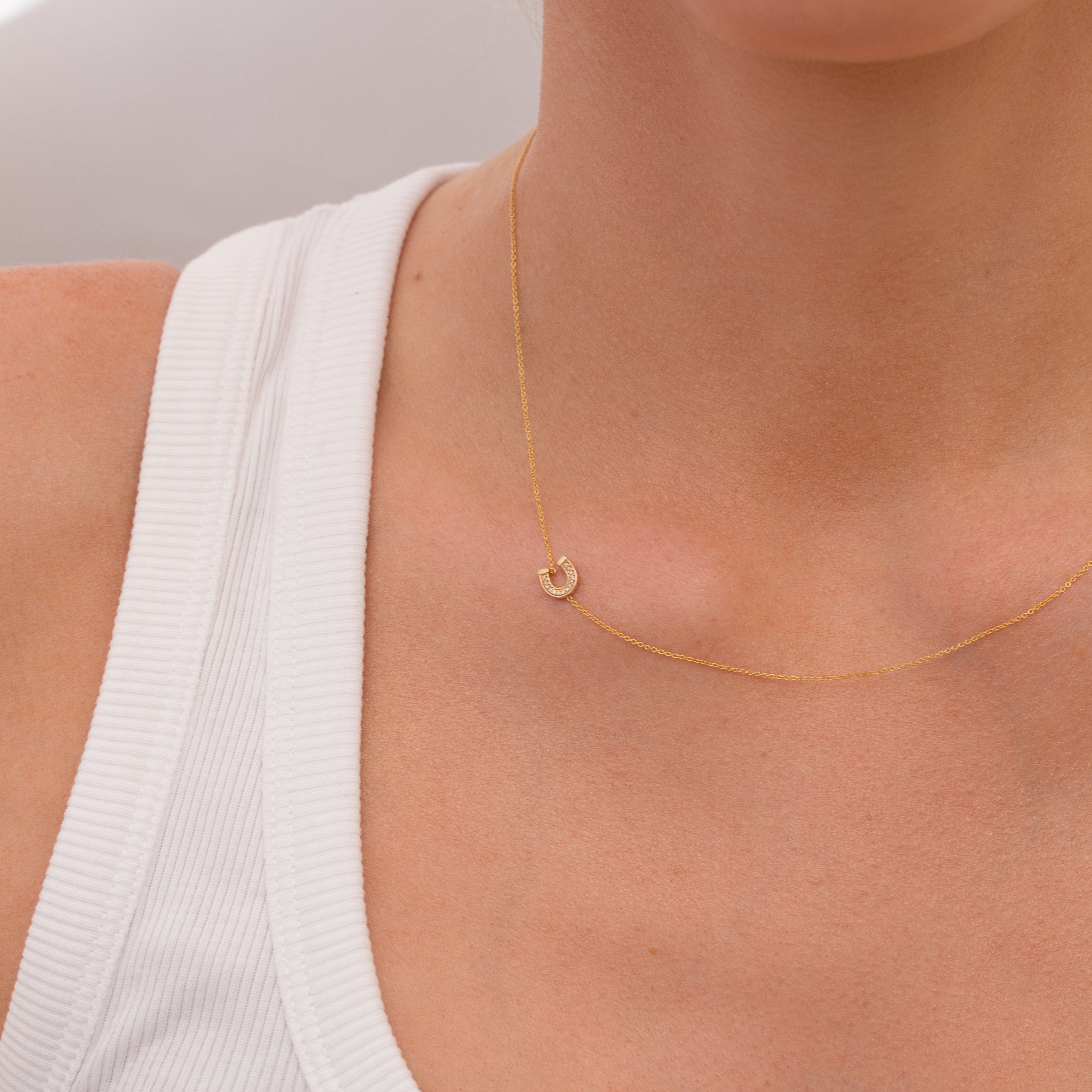 Gold necklace with a small pavé diamond horseshoe charm on a person wearing a white top against a neutral background
