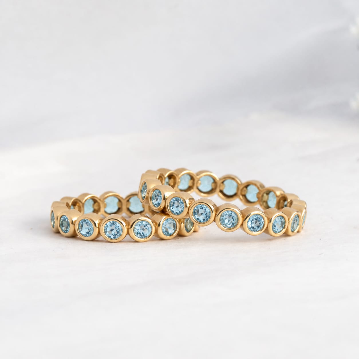 A gold eternity ring featuring round-cut light blue gemstones in a bezel setting, photographed on a soft white marble surface.