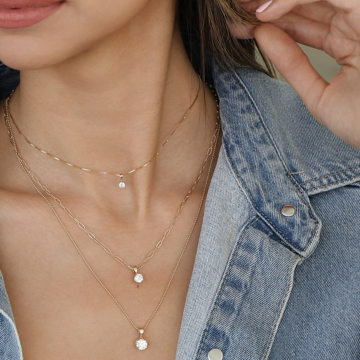 Close-up of a woman's decellotage wearing a Duchess Diamond Pendant on The Slide Chain, a Princess Diamond Pendant on an Element Short Link Chain and a Queen Diamond Pendant on a Bead Chain, all in yellow gold, with a denim jacket. 