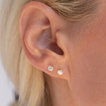 Close-up of a person's ear wearing two small round stud earrings with clear sparkling stones in a silver-toned setting.
