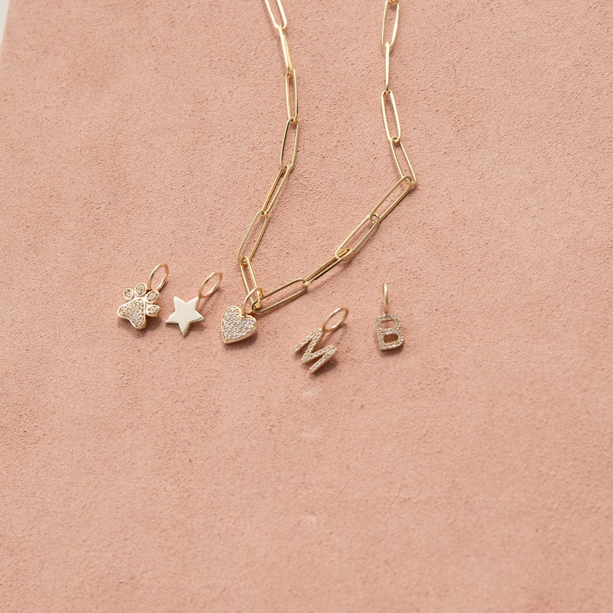 An Element Long Link Chain with Charm Pendants Star, Pave Paw and Pave Heart, and Pave Letter Charms in M and B, against a light pink background. 